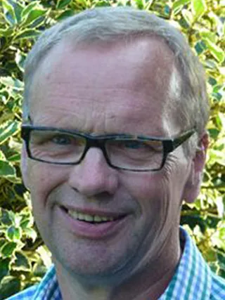 Porträt eines lächelnden Mannes mit Brille vor grünem Laub.