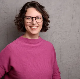 Porträt einer lächelnden Frau mit Brille und schulterlangem braunem Haar im Freien.
