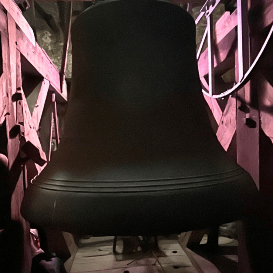 Massive Kirchenglocke mit rosa Licht beleuchtet, im hölzernen Glockenturmgerüst.