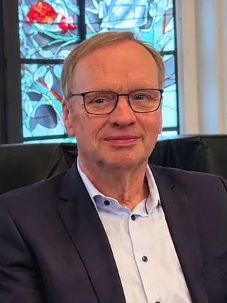 Porträt eines lächelnden Mannes mit Brille vor grünem Laub.
