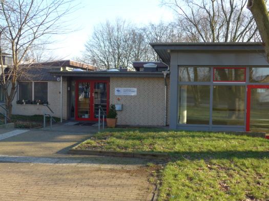 Eingang eines Backstein-Gemeindezentrums mit großen Fenstern und roter Tür.