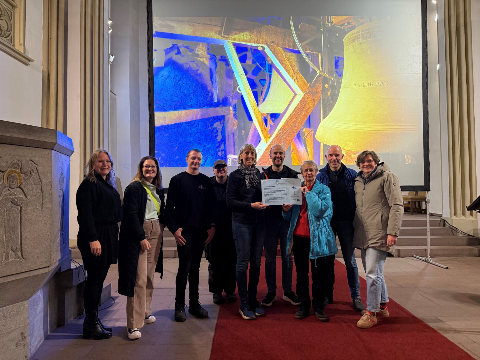 Gruppe hält UNESCO-Zertifikat in Kirche vor Projektion von Kirchenglocken.