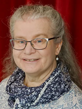 Porträt einer lächelnden Frau mit langen blonden Haaren, Brille und Perlenohrringen.