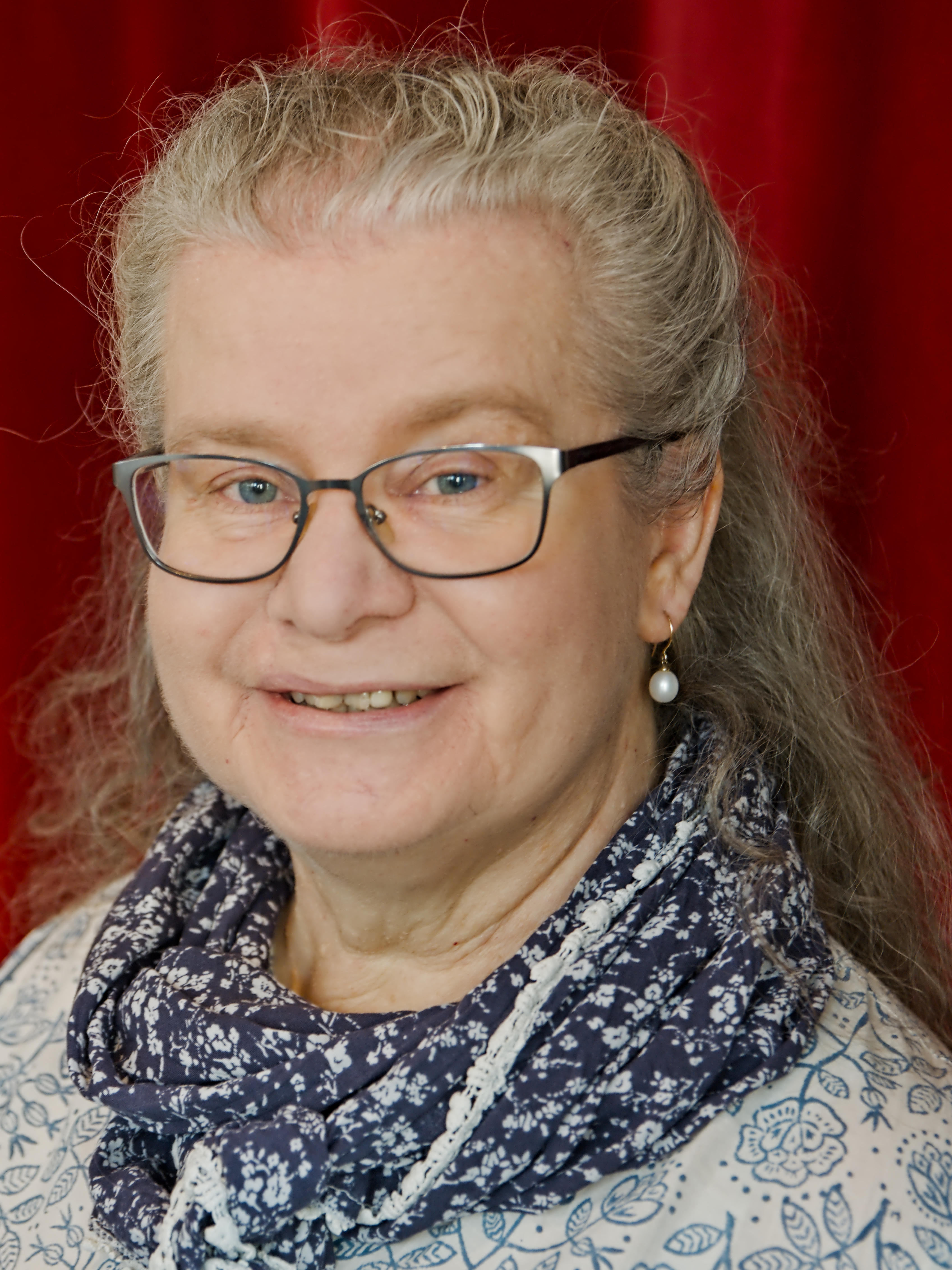Porträt einer lächelnden Frau mit langen blonden Haaren, Brille und Perlenohrringen.