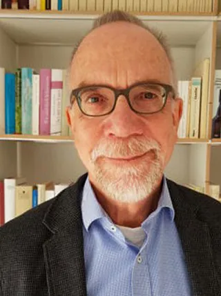 Porträt eines lächelnden älteren Mannes mit Brille und Bart vor einem Bücherregal.