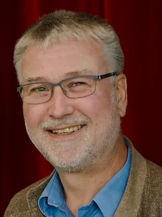 Lächelnder Mann mit Brille und grauem Haar lehnt an dunkler Holzfläche in einer Kirche.