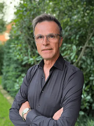 Porträt eines Mannes mit Brille, trägt schwarzes Hemd, Arme verschränkt, steht im Freien.