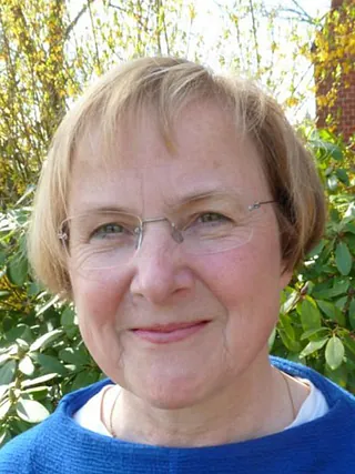 Porträt einer lächelnden Frau mit kurzen blonden Haaren und randloser Brille im Freien.