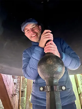 Mann lächelt unter einer Kirchenglocke und hält den Klöppel.