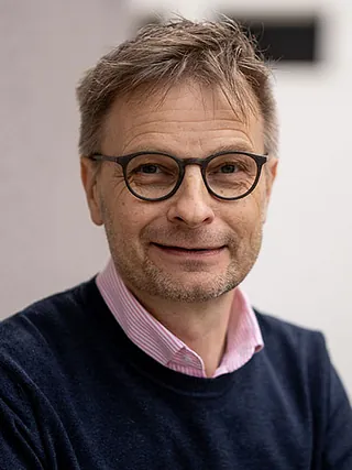 Porträt eines lächelnden Mannes mit Brille, trägt blauen Pullover und rosa gestreiftes Hemd.